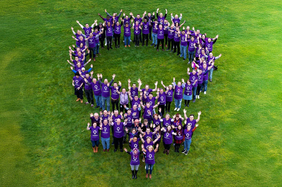 Group of people standing in a shape on grass and waving