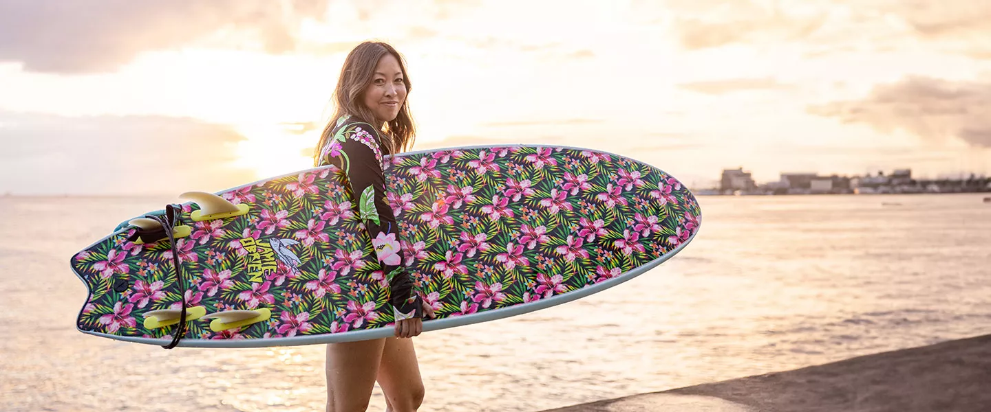 Woman holding surfboard and walking on the beach in front of a sunset.